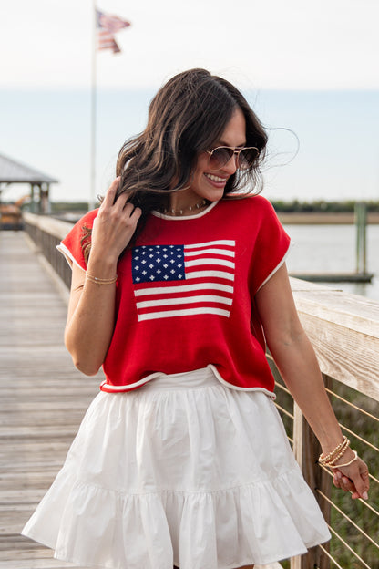 Woman wearing a red shirt with an American flag design and a white skirt on a wooden dock.