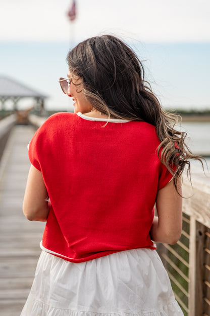 Woman in a red shirt and white skirt standing on a wooden pier by a body of water.