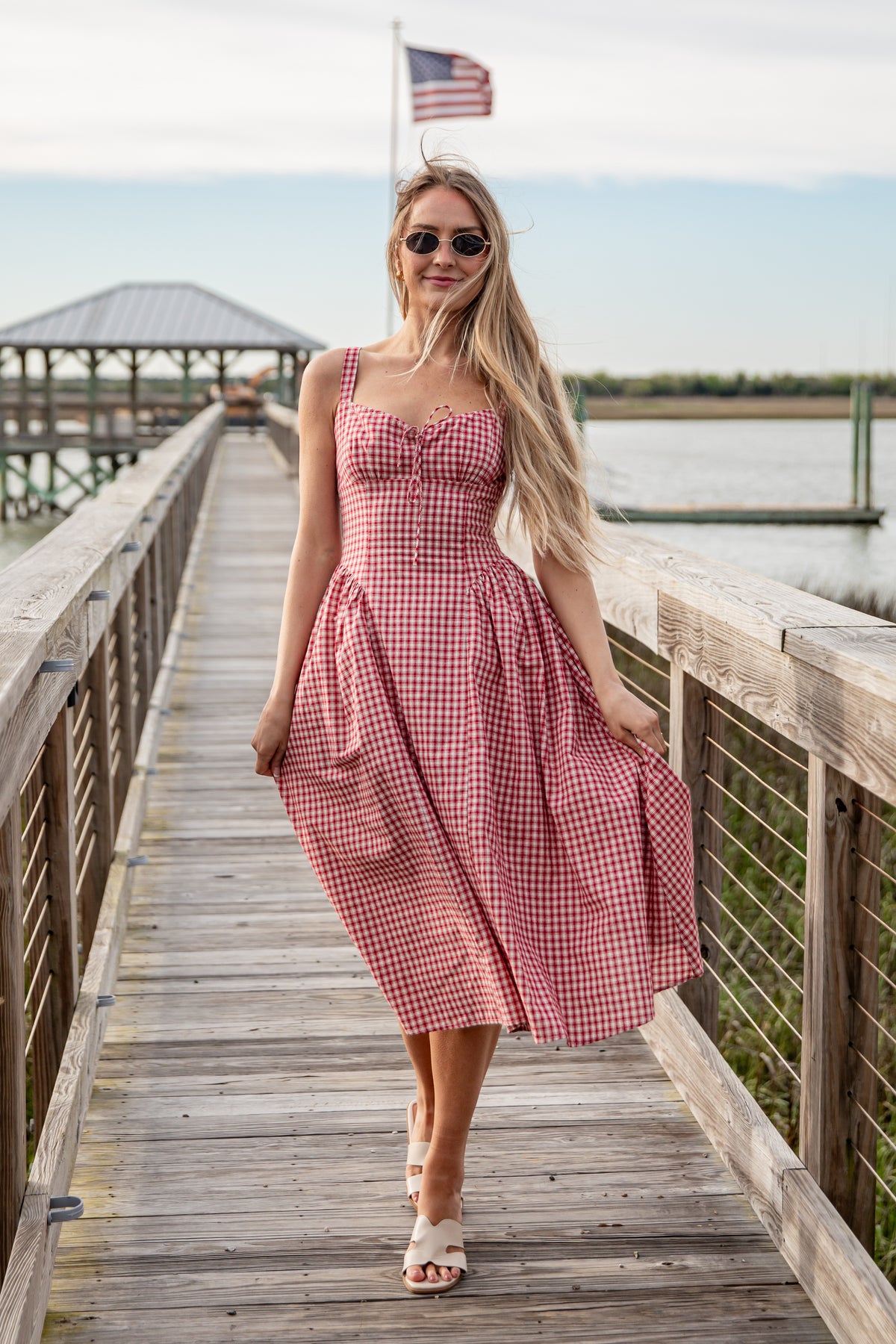 Woman in a red checkered dress standing on a wooden dock with an American flag in the background.