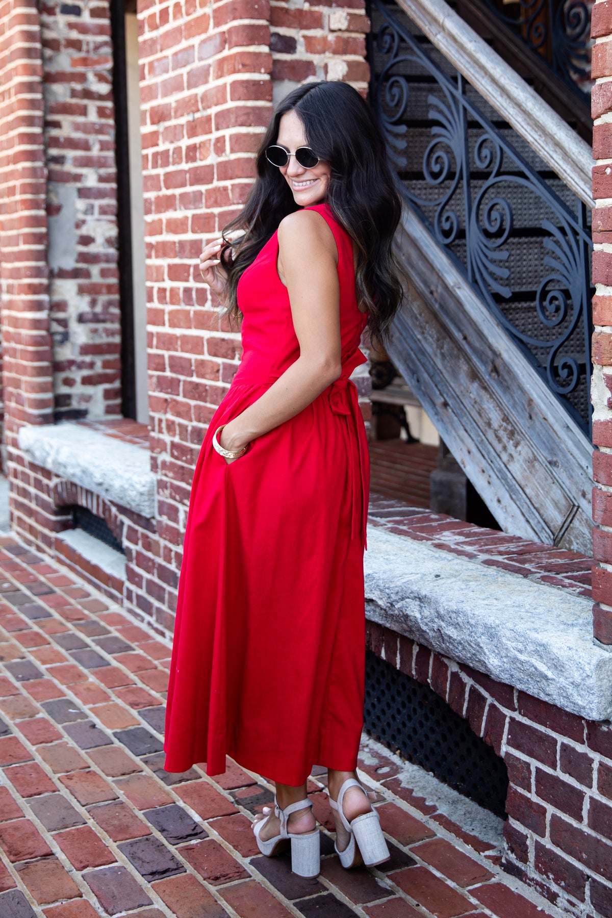 A woman wearing a sleeveless red midi dress and white platform sandals stands on a brick sidewalk beside a red brick building with a staircase.