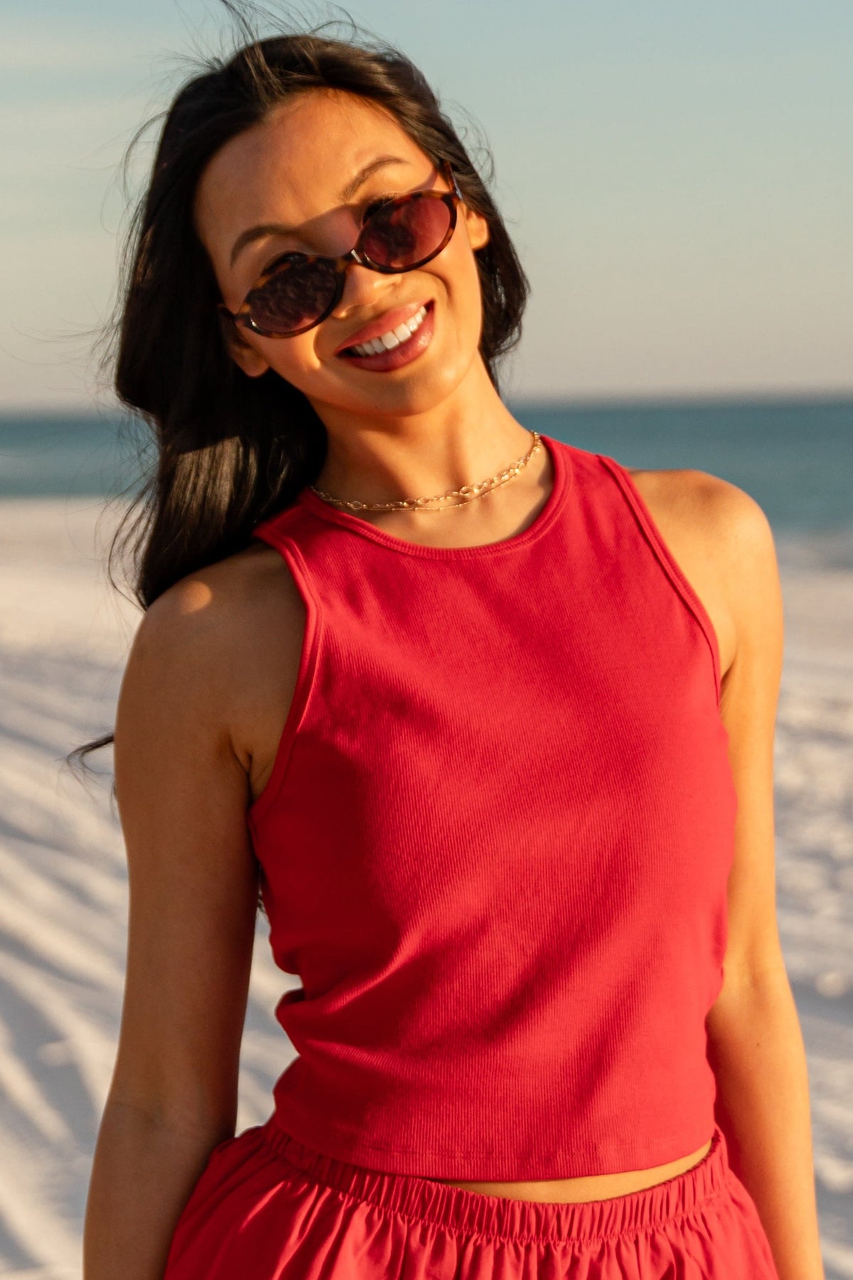 Woman in a red outfit standing on a sandy beach with sunglasses.
