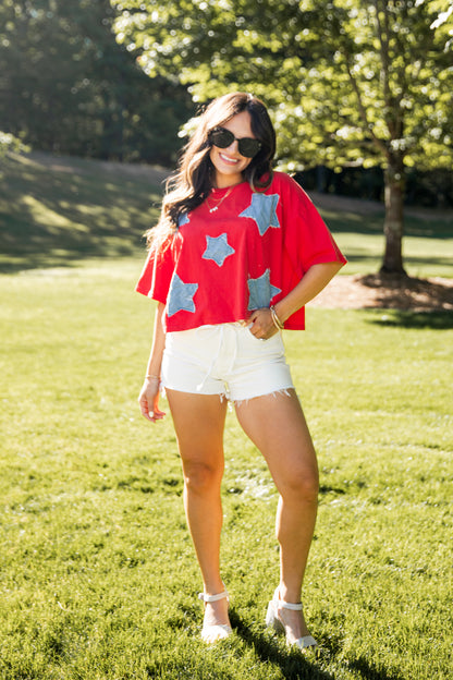 Woman wearing a red shirt with blue star patterns and white shorts standing on grass.