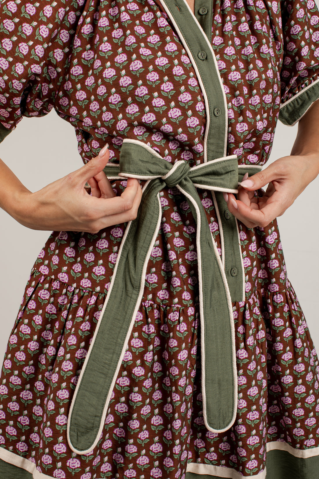 Close-up of a person wearing a brown dress with a pink floral print and an olive green self-tie belt with cream trim, adjusting the belt at the waist.