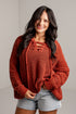 A woman wearing a rust-orange chunky knit lace-up sweater and light-wash denim shorts, standing against a light gray wall.