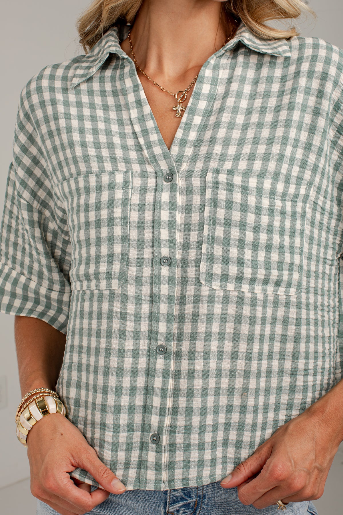 A woman wearing a green and white gingham check short-sleeve button-up shirt with two chest pockets, paired with blue jeans and gold jewelry.