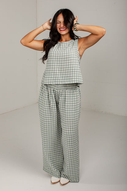 A woman modeling a matching two-piece lounge set featuring a sleeveless crop top and wide-leg pants in a sage green and white gingham check pattern, set against a plain light gray studio background.