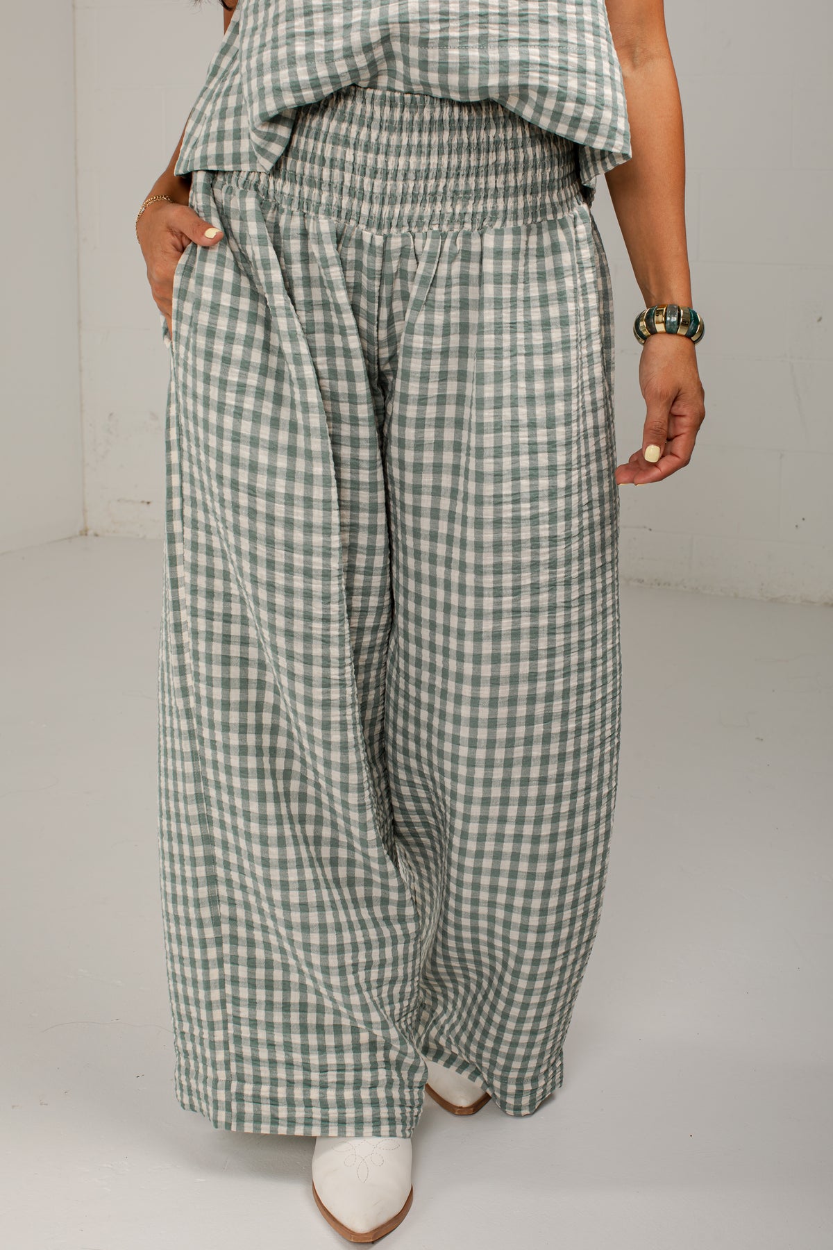 A person wearing wide-leg cropped pants in a green and white gingham check pattern, paired with a matching green and white gingham top and white pointed-toe heels, photographed against a plain light gray studio background.