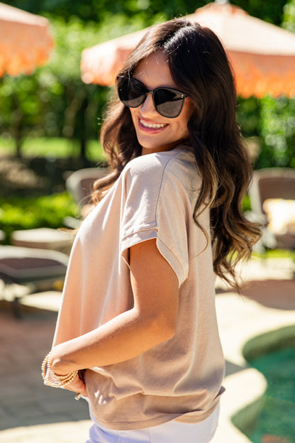 Woman wearing sunglasses and a beige top in an outdoor setting with orange umbrellas.