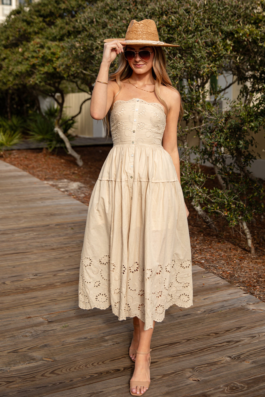 Woman in a beige dress and hat standing on a wooden boardwalk with greenery and a house in the background.