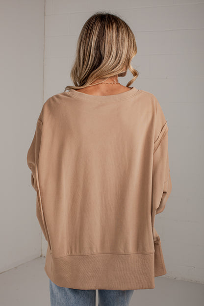 A woman viewed from behind wearing a loose, oversized tan crewneck sweatshirt and light blue jeans, standing against a white paneled wall.