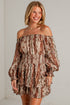 A woman modeling an off-the-shoulder mini dress with a brown and beige paisley pattern, paired with beige ankle boots, standing against a white brick wall.