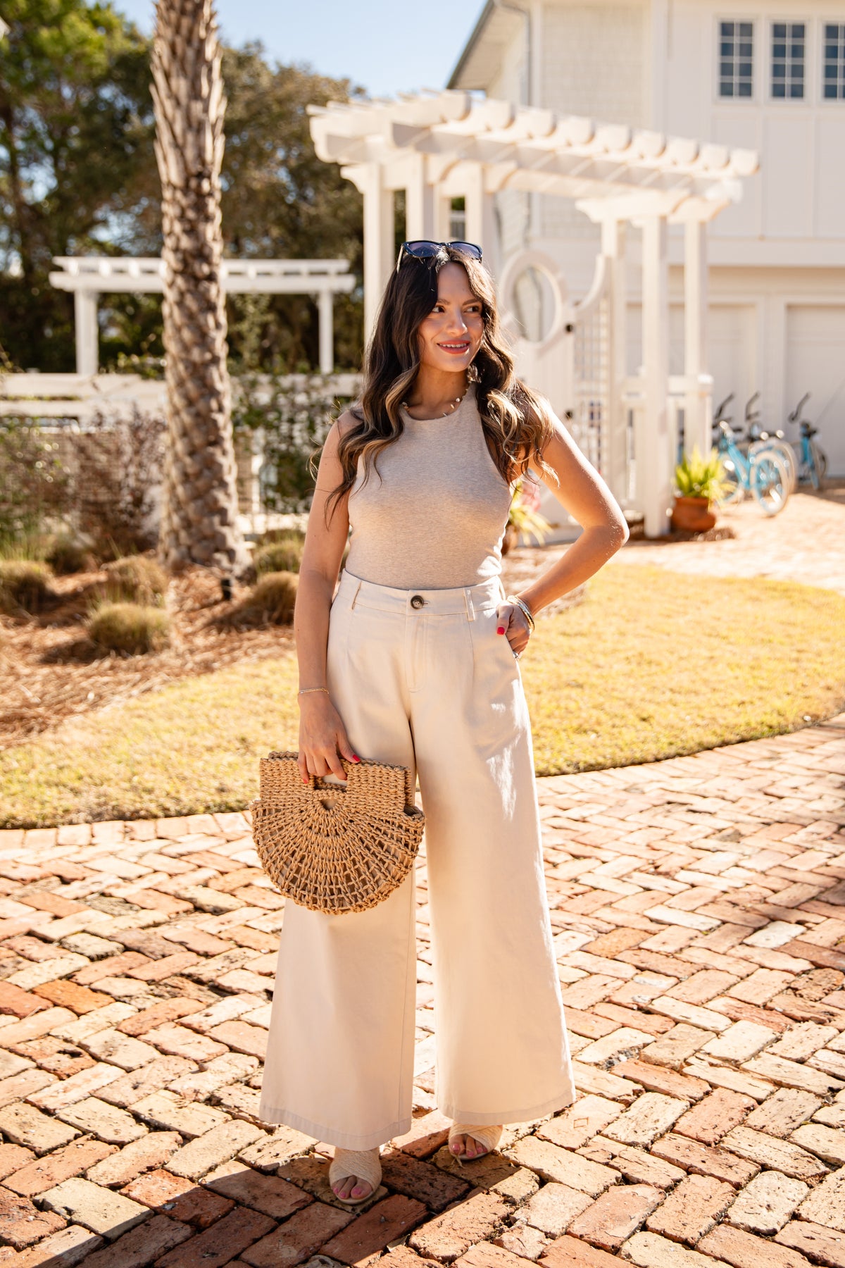 Woman in a beige top and white pants standing on a brick path with a white building and trees in the background.
