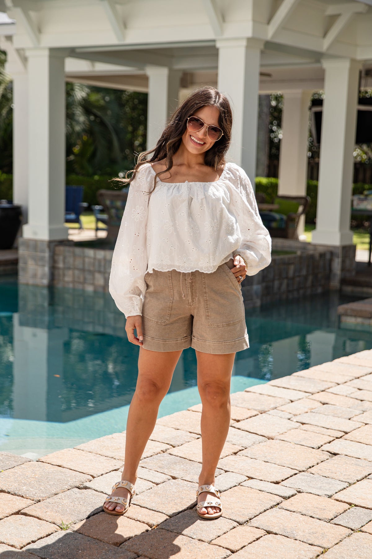 Woman standing by a pool wearing a white blouse and beige shorts.