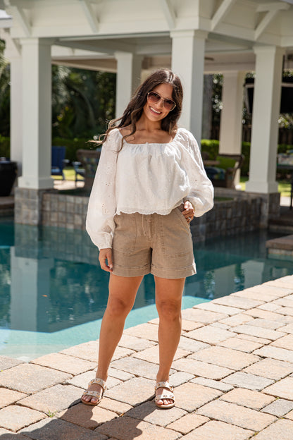 Woman standing by a pool wearing a white blouse and beige shorts.