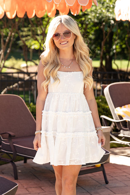 Woman in a white dress standing outdoors with chairs and greenery in the background