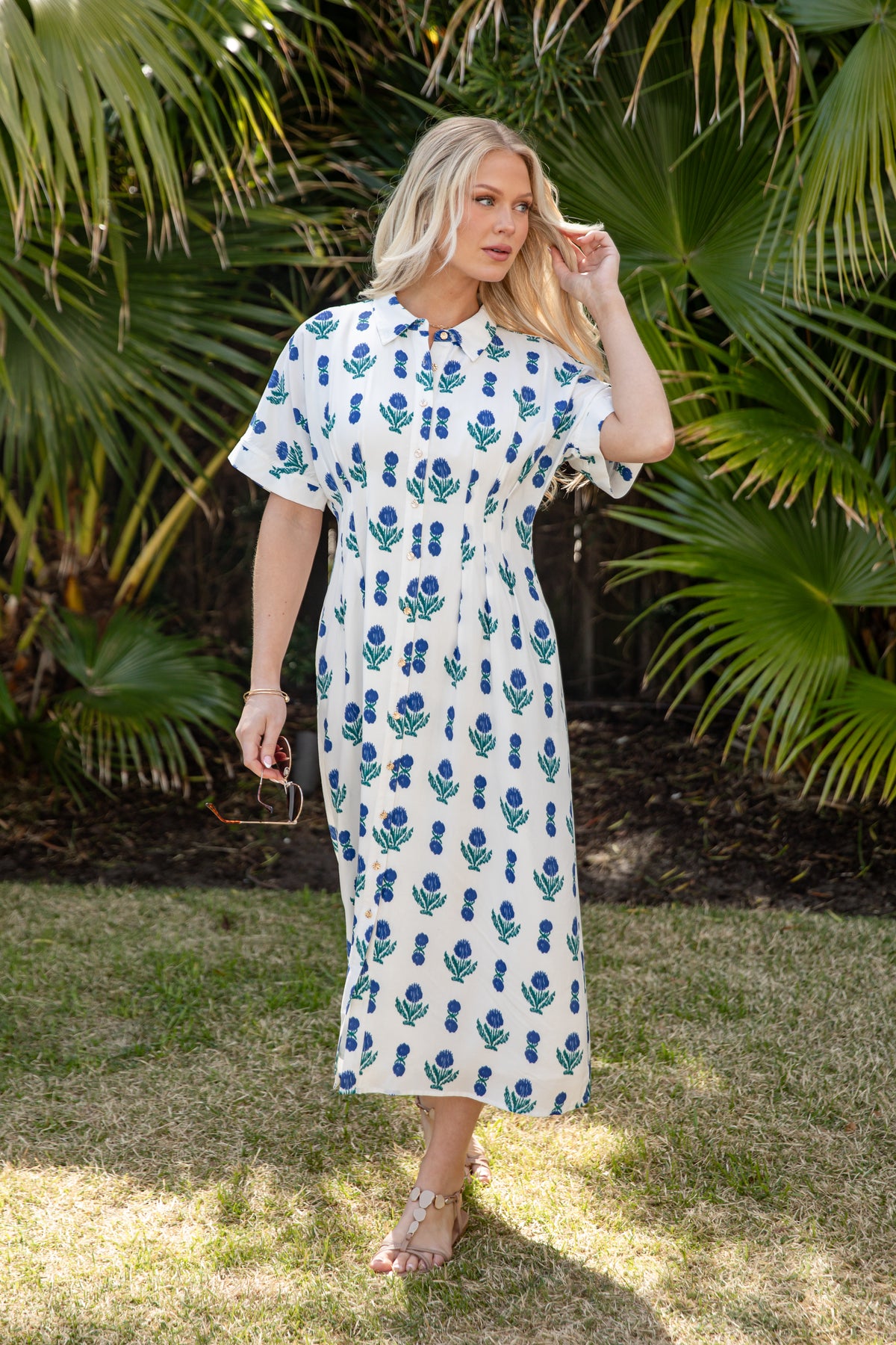Woman in a white dress with blue floral pattern standing outdoors with greenery in the background