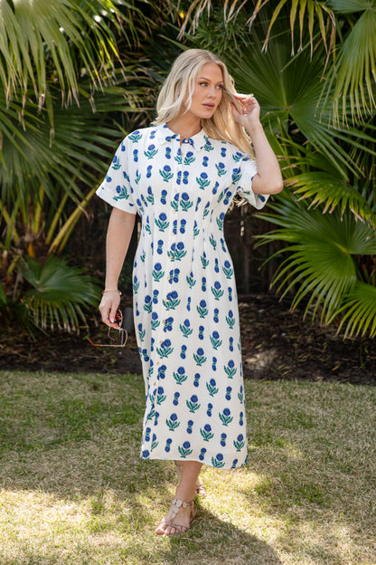 Woman in a white dress with blue floral pattern standing outdoors with greenery in the background