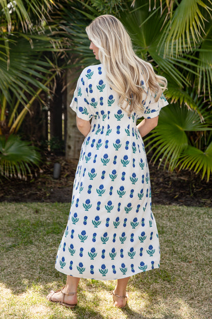 Woman wearing a white dress with blue floral pattern standing in a garden.