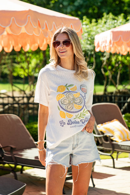 Woman wearing a graphic t-shirt and denim shorts standing outdoors with lounge chairs and umbrellas in the background.
