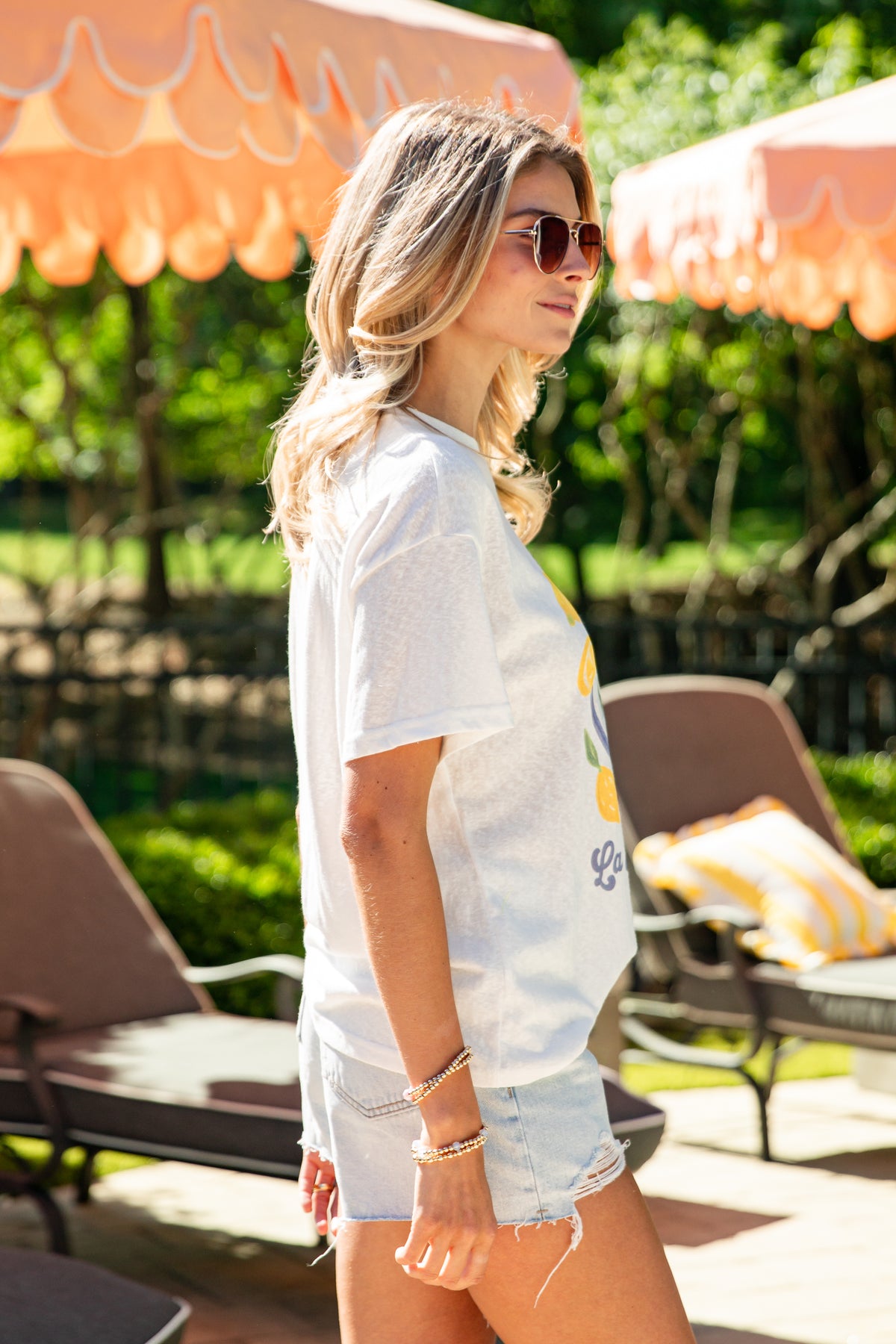 Woman in a white t-shirt and denim shorts standing in a garden setting with orange umbrellas and lounge chairs.