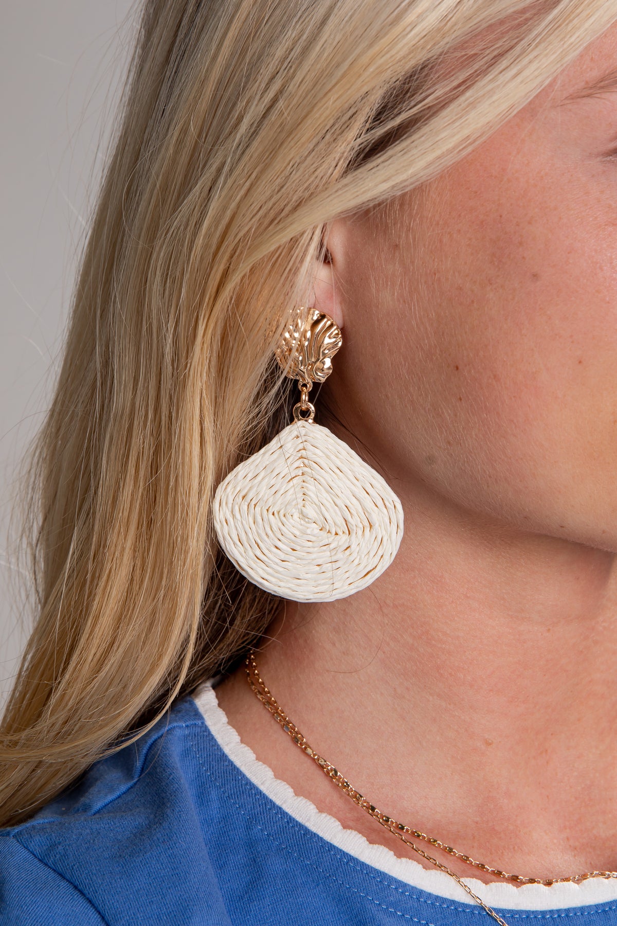 Close-up of a person wearing beige raffia earrings with a blue shirt and necklace.