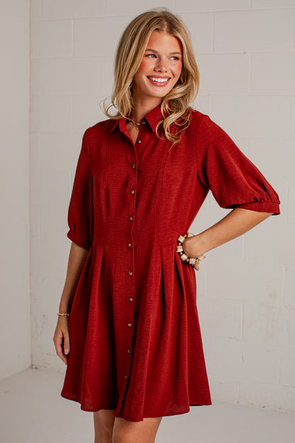 A woman modeling a red button-down shirt dress with puff sleeves and a flared skirt, standing against a white brick wall.