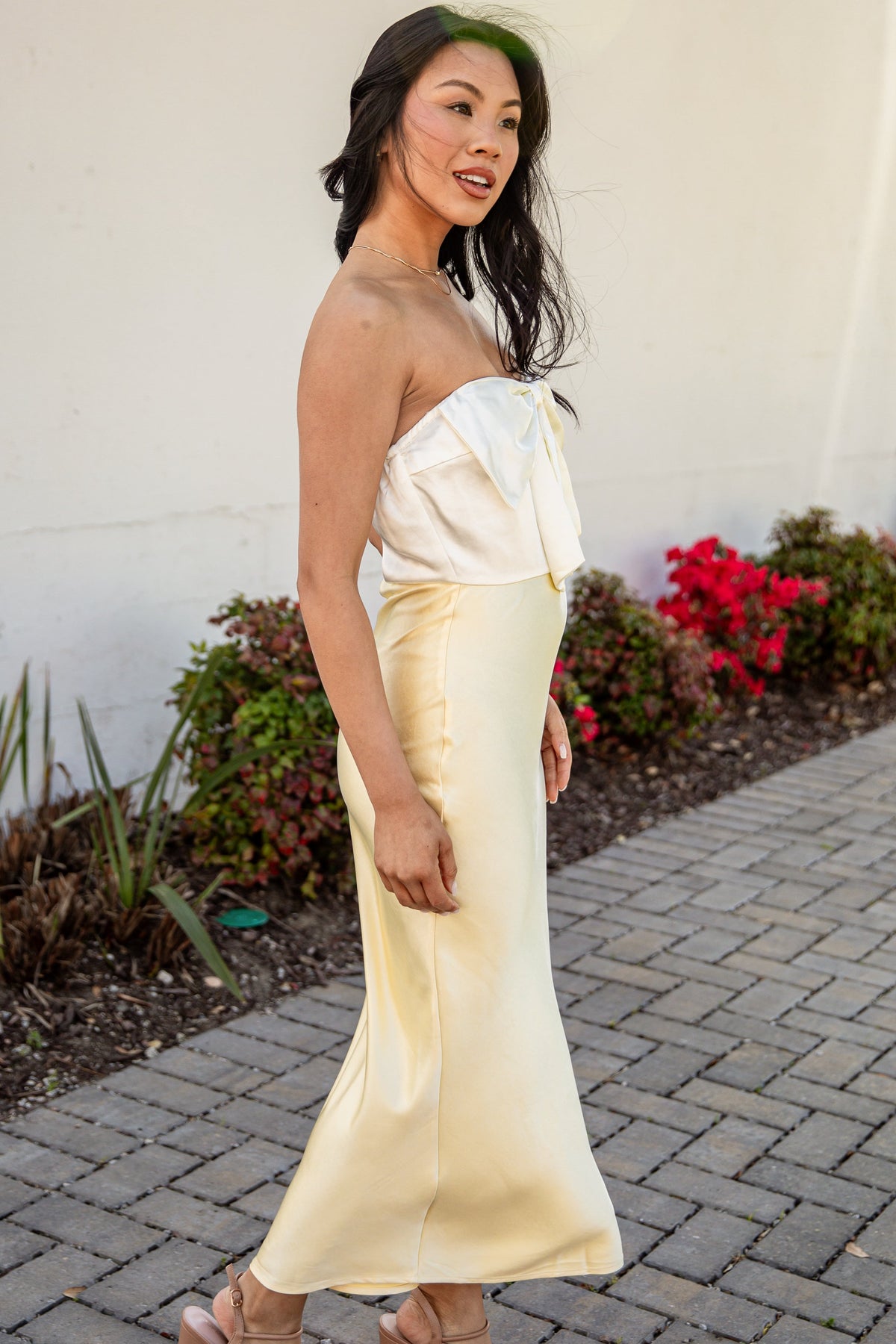 Woman in a strapless yellow dress standing on a sidewalk with plants in the background