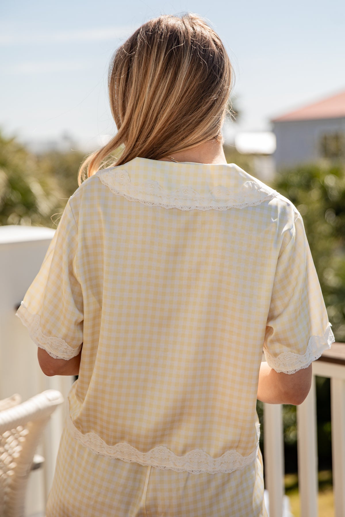 Woman wearing a light yellow checkered shirt on a balcony with greenery and a clear sky.