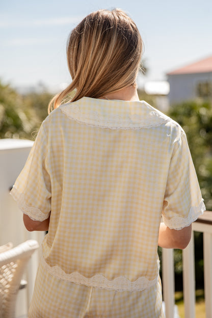 Woman wearing a light yellow checkered shirt on a balcony with greenery and a clear sky.