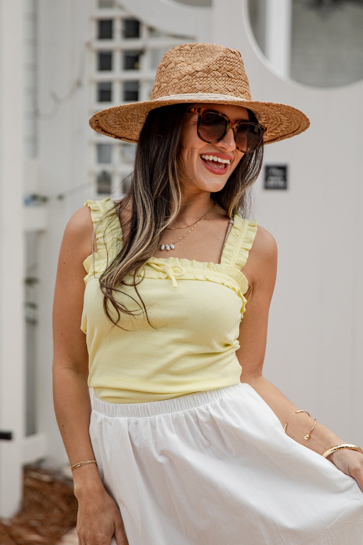 Woman wearing a yellow top and white skirt with a straw hat and sunglasses, standing against a blurred outdoor background.